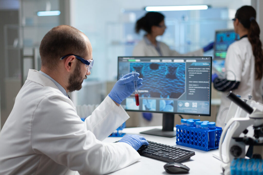 : Scientist examining a test tube while analyzing data on a digital monitor.